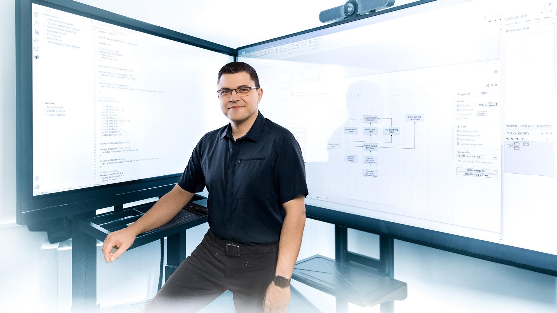 Hendrik steht vor zwei großen Bildschirmen, die technische Diagramme und Code anzeigen.