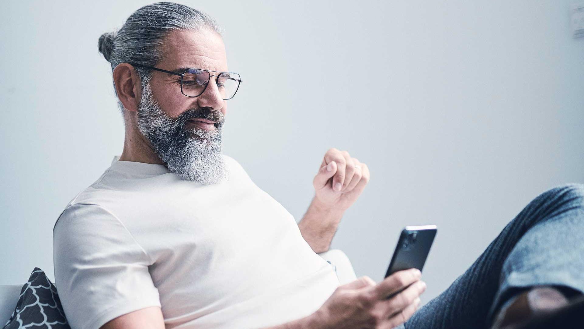 A man with gray hair, a full beard and glasses sits on the couch and looks at a smartphone
